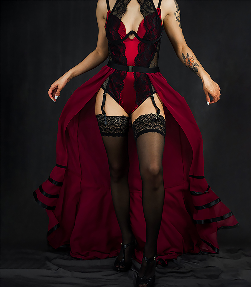A full front view of this glamorous red bodysuit is made of silk charmeuse, black lace, and dotted mesh. The bodysuit features a keyhole neckline with a high-neck lace overlay. It is paired with a red chiffon skirt that has ruffles at the bottom and finished with black satin ribbon.