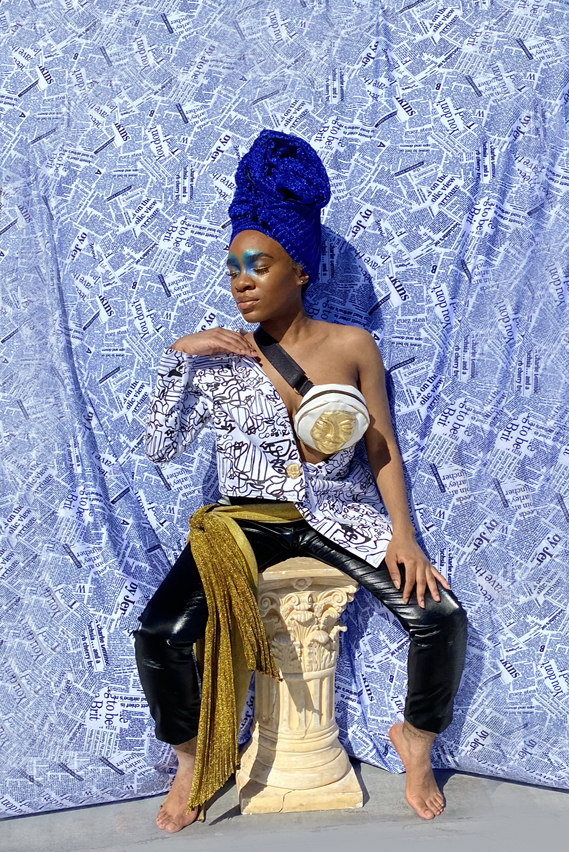Woman seated on a column in shimmer blue head wrap. Abstract black and white print asymmetrical blazer. Black faux leather pants with golden glitter waist sash. Newsprint backdrop. Golden mask fanny pack accessory. Dramatic blue eyeshadow on inner eyelid and inner eyebrows.