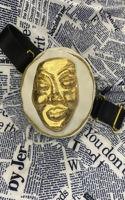 White leather round fanny pack with gold hardware details and black straps. Golden face mask detail placed in front.