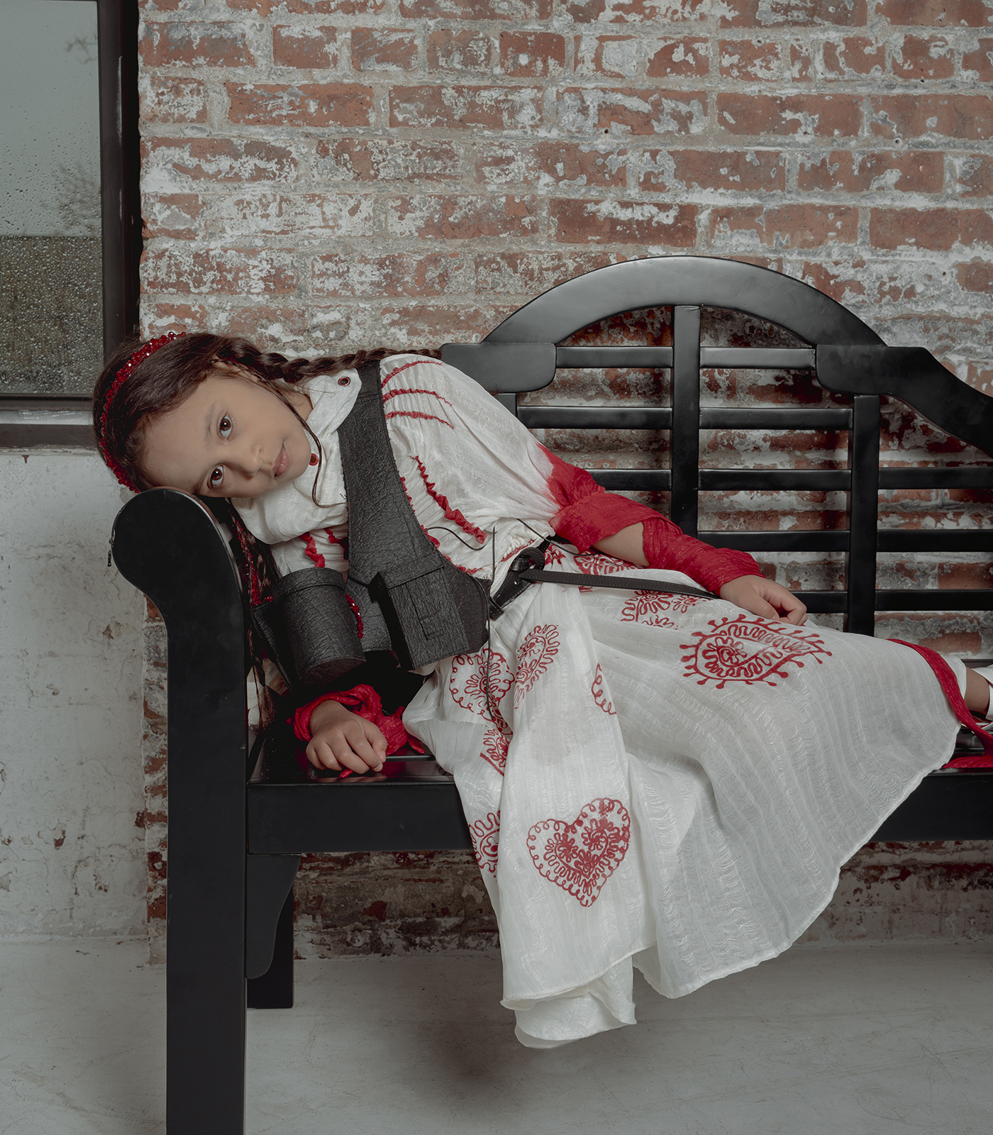 An eight-year-old brown hair girl leaning on a black bench wearing a high neck white silky blouse and a red heart pattern printed box pleat skirt and a black vest, a background of brick wall and rainy windows.