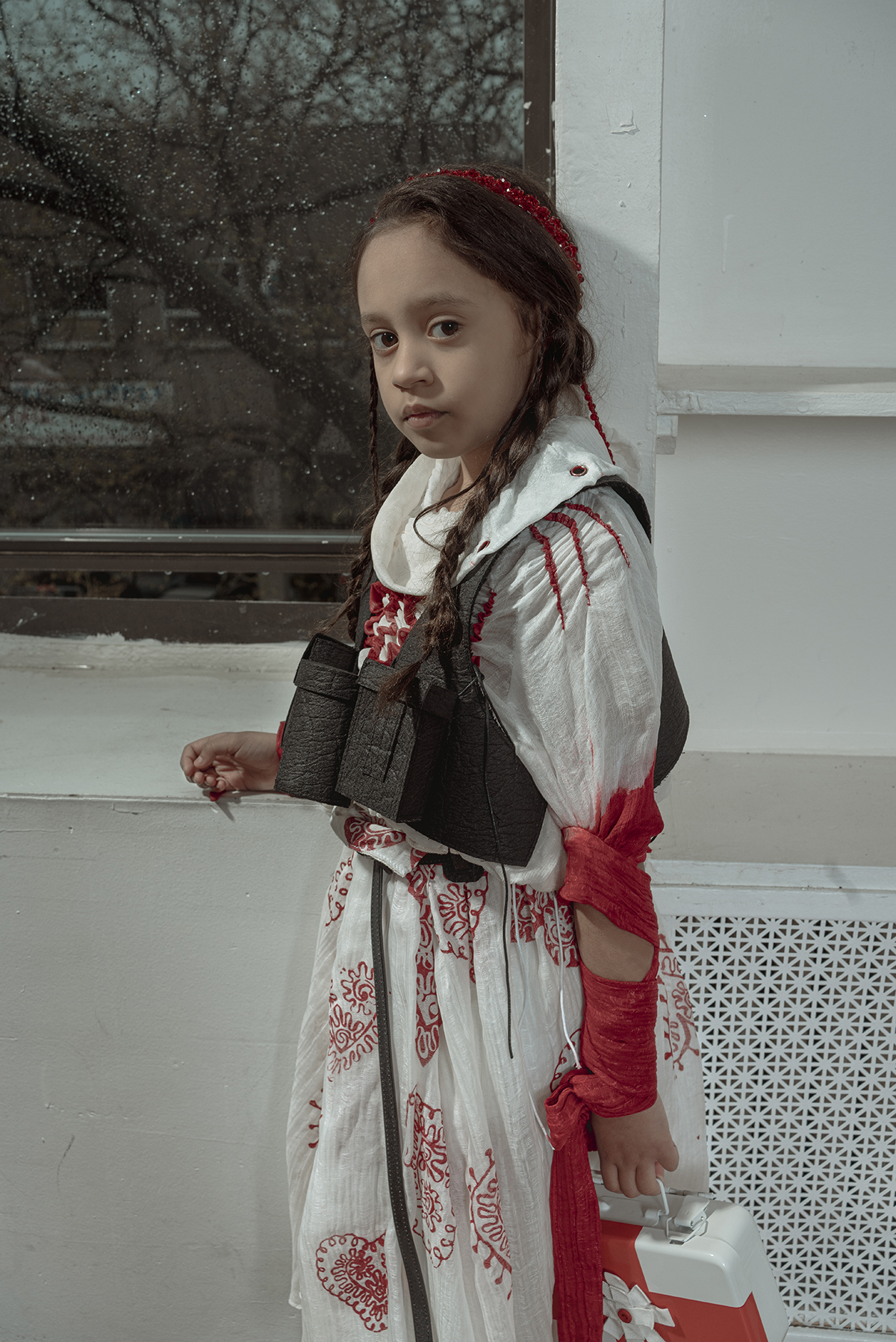 An eight-year-old brown hair girl holding a first-aid box, wearing a turtle neck white silky blouse, a red heart pattern printed box pleat skirt and a black vest, standing aside of a rainy window.
