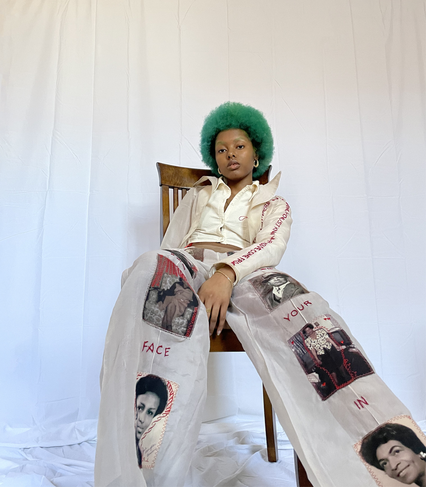Person sitting in chair. Clothing: Beige silk chiffon blazer and pant and a cropped cotton seersucker button up. Blazer is hand embroidered and states "To know where you're going you must know where you've come from." Pant has personal family photos stitched/patched onto the font of the pant leg. In addition, hand embroidery states "I look for your face in my reflection."