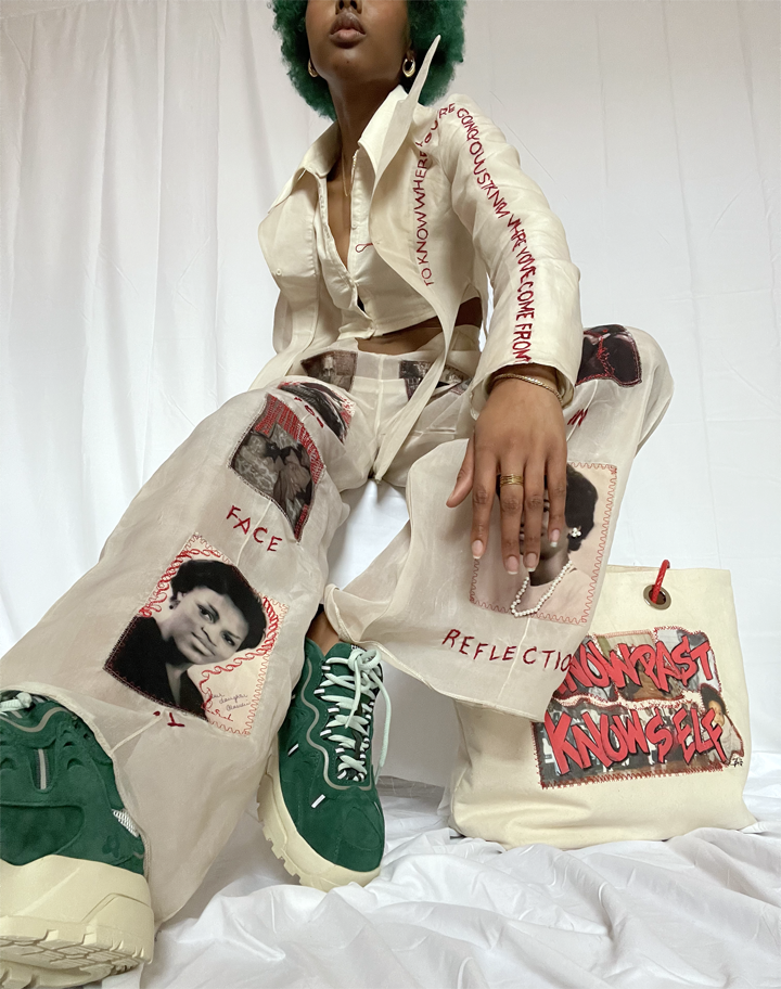 Young woman sitting on chair with a bag beside her. Clothing: unbuttoned beige blazer revealing a ivory seersucker cropped button up underneath. Pants are beige with portrait photos sewn on top of the pant with accents of red embroidery.