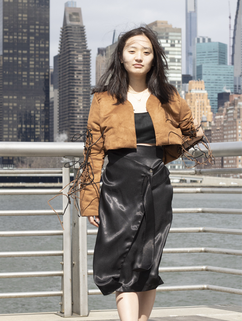 Black and brown suede cord trim application on brown suede cropped jacket enhanced by wind. Fluidity/movement of black crepe back satin skirt also shown in wind. Skirt has a wrap feature (coming from the model's right side seam of the skirt) where it is snapped on in place and has a tie feature (coming from the model's left side seam of the skirt), which goes through the opening slot that is on the wrap skirt feature part of the skirt.
