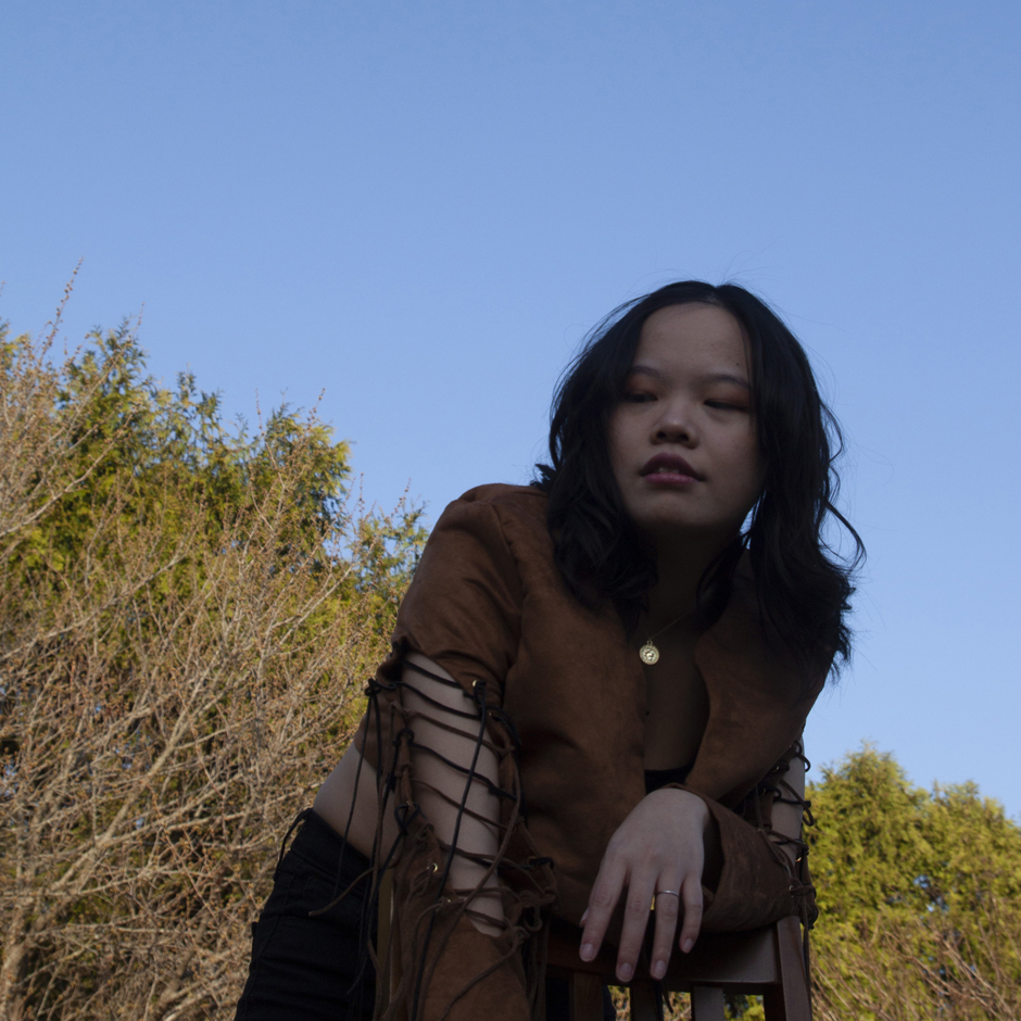 Model leaning on chair exposing the sleeve cutout and fringe of the brown suede jacket.