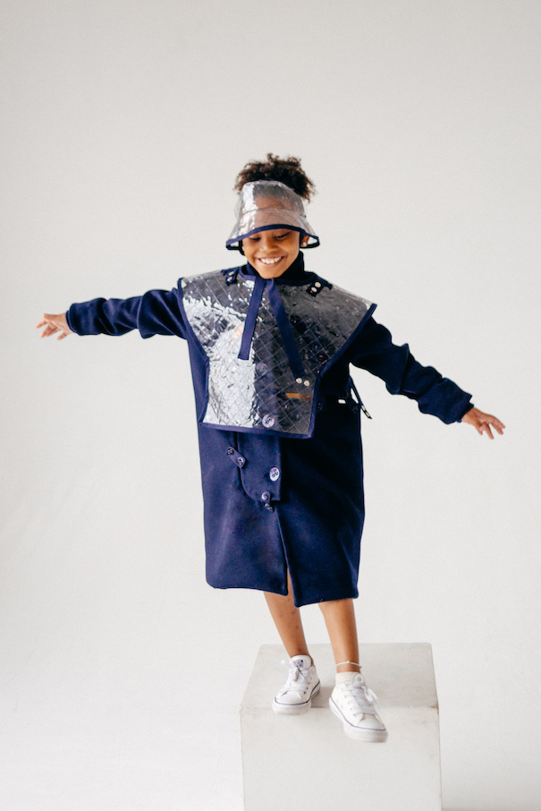 The model is wearing a patched wool trench coat with a quilted vinyl poncho and hat layered on top.
