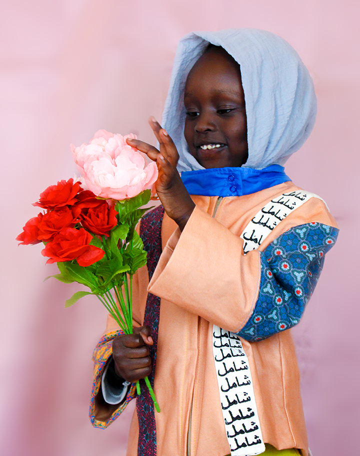 The child model is holding silk flowers that are red and pink while smiling. The Arabic on the white strap of the jacket reads “Inclusive”.