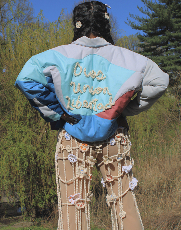 With her hands on her hips, the model shows the back of the jacket. The back of this jacket also has geometric shapes as well as the words "Dios, Union, Libertad" crocheted on which is the motto of El Salvador, which means "God, Union, and Liberty."