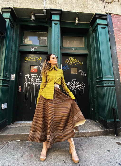 A front view of the look without the bomber jacket. The model is playing with the sweep and movement of the skirt, displaying the contrast denim hem facing. The taffeta blouse is now visible, displaying the exaggerated collar and extra large cuff. The background is the same as the previous images.