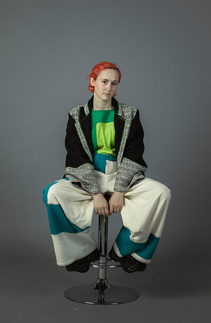 Photo of a girl seated on a stool. Her legs are bent and point outward in a V-shape. Her feet rest on the footrest about a foot above the floor, and her large, chunky shoes are visible below the hemline of her pants. She has her hands placed on the stool in between her legs. Her right shoulder is tilted slightly down and her head tilts toward her left shoulder. She is wearing a loose-fitting green top, white and blue wide-leg pants, and a black loose-fit jacket that is open. Her hair is red, and she is looking at the camera.