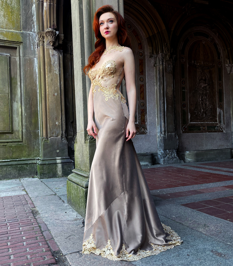 A woman wearing a taupe long silk charmeuse gown with gilded embroidered net appliqué hand-sewn on a sheer net bodice with beaded embellishment. She is standing in front of an old, magnificent arch.