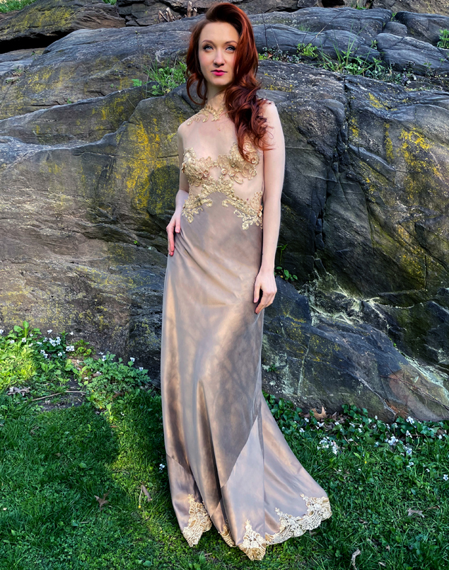 The woman smile slighlty and is facing us, standing on the green grass with grey rocks and nature behind her. She is wearing a taupe long silk charmeuse gown with gilded embroidered net appliqué hand-sewn on a sheer net bodice with beaded embellishment. 