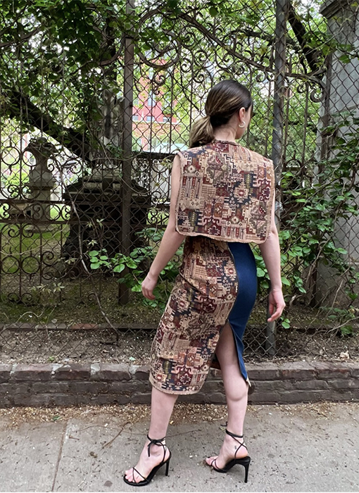 On the road in front of the park, the model 's back is to the park, her face is turned to the camera, her hands are folded in front, and her head is turned to the right. The garden and statues are visible behind the wires.