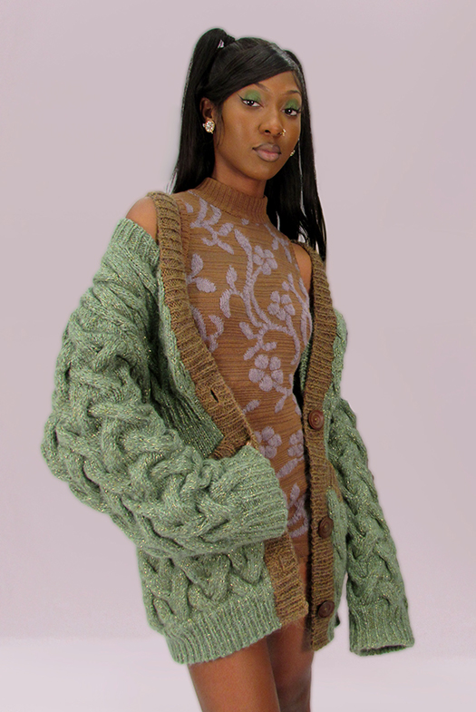 This cardigan is made from a melange of yarns, including mohair, lambswool/silk, and metallic yarn. It is fern green with specs of gold throughout. Image of a girl wearing cardigan off the shoulder and open revealing bodysuit. Bodysuit has large floral design.The cardigan is hand-knit with medium-sized lattice cables. The background is a warm grey.