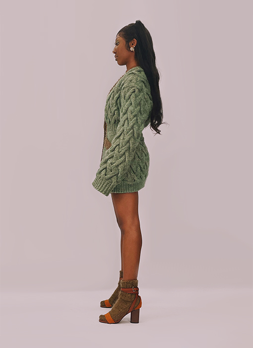 This cardigan is made from a melange of yarns, including mohair, lambswool/silk, and metallic yarn. It is fern green with specs of gold throughout. We see a girl's full side profile showing off the cable sleeves of the cardigan..The cardigan is hand-knit with medium-sized lattice cables. The background is a warm grey.