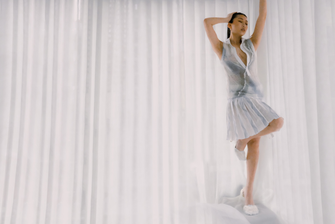 A woman stands to the right of the composition with her arms raised above her head as she closes her eyes. A sleeveless top with blue and teal dyes is combined with a pleated skirt of similar color orientation. The woman is posed on one leg, with the other raised and bent like a flamingo, highlighting white and blue fluffy mohair heeled sandals.