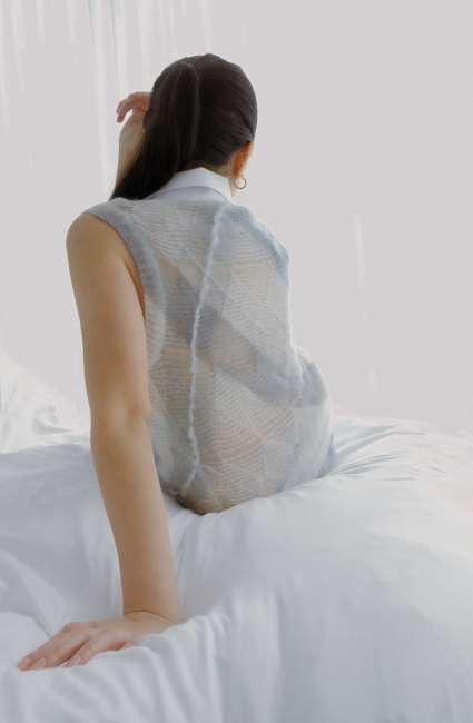 A woman sits with her back facing the viewer, hand posed behind her as her support. Clearer detail of mohair embroidery and stitch-transferred diamond patterns are visible, as well as the diagonal, thick stripes of light blue dye.
