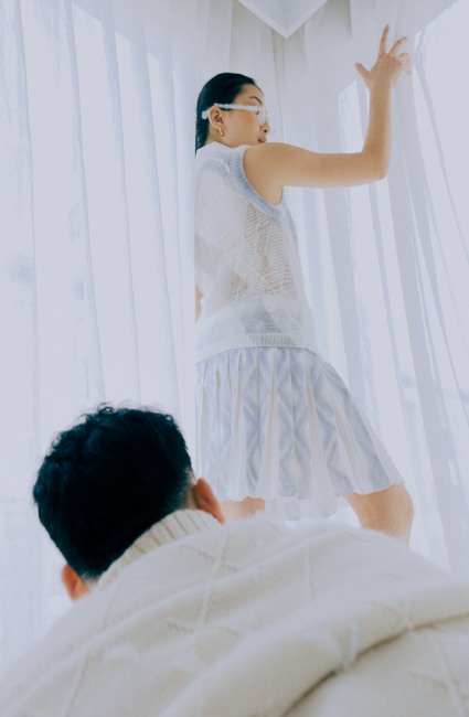 A man wearing a thick beige jacket with partially rendered Aran diamonds gazes up at a woman wearing a sleeveless top and pleated skirt. Both face away from the viewer as the woman puts a hand on the curtain.
