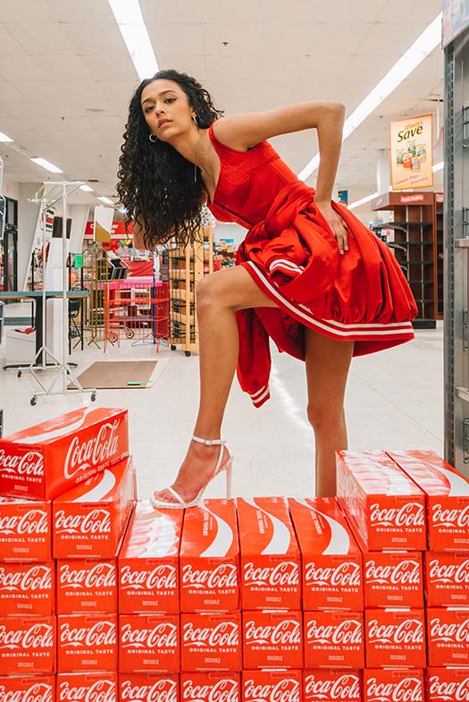 Model poses with Coca-Cola 12-pack cans with one leg up on the packs.