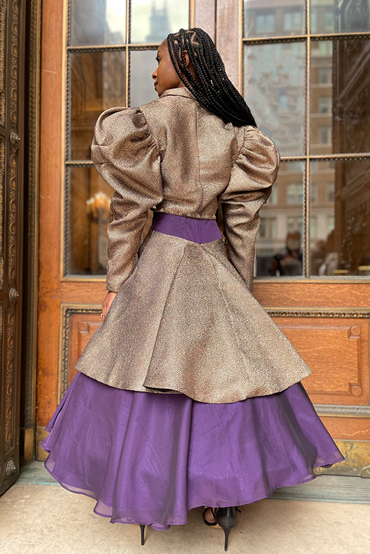 A back view of the tea-length dress and the jacket. The jacket sleeves are detailed with two darts near the top cap of the sleeves, alongside tucks that were created and sewn around the sleeve's seam allowance. The pearl beading is continued around the back of the cropped jacket hemline, which shows off the peaking of the plum purple dress before the eye cascades down to the layered skirts. The top black-and-gold metallic skirt naturally flares in the back, giving a sense of drama.