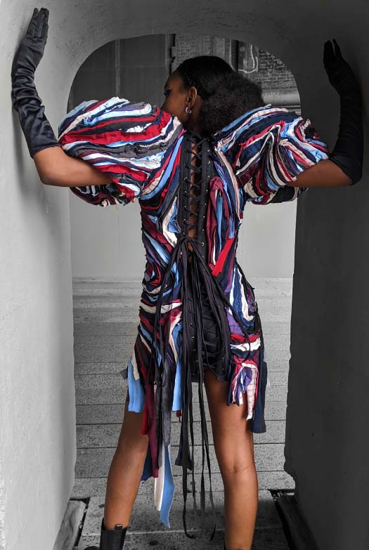 The model stands with her back toward the camera as she enters a small rounded doorway.  She places her hands up to the top of either side of the doorway and looks off to the left.  This view displays the 19th-century corset construction of the garment through the center back lacing panel, which extends from the top of the neck down to the miniskirt-length hem. The model is also wearing a pair of black spandex shorts underneath the corseted dress.
