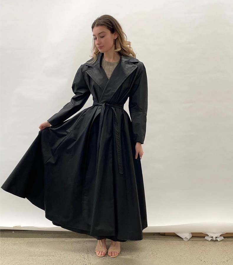 Girl holding long trenchcoat off to one side. The trenchcoat is made out of faux black leather, has a large notch collar, and is belted at the waist.