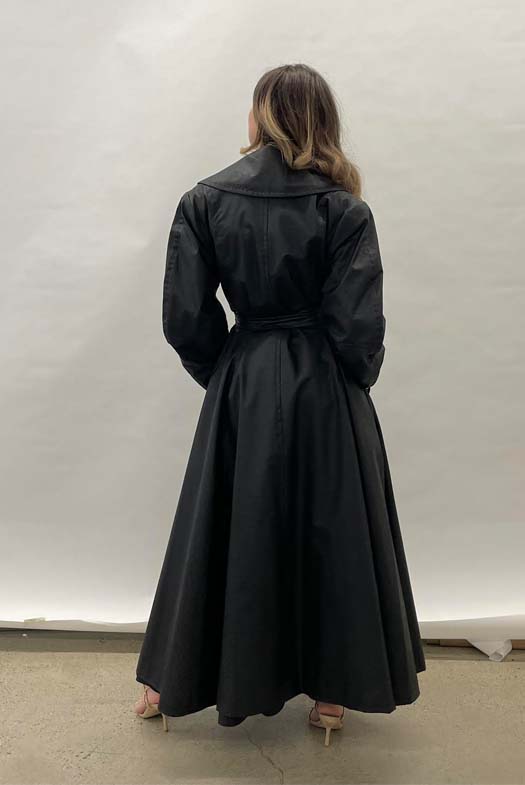 Back view of a girl wearing long trenchcoat made out of faux black leather and has a large notch collar and belted at the waist.
