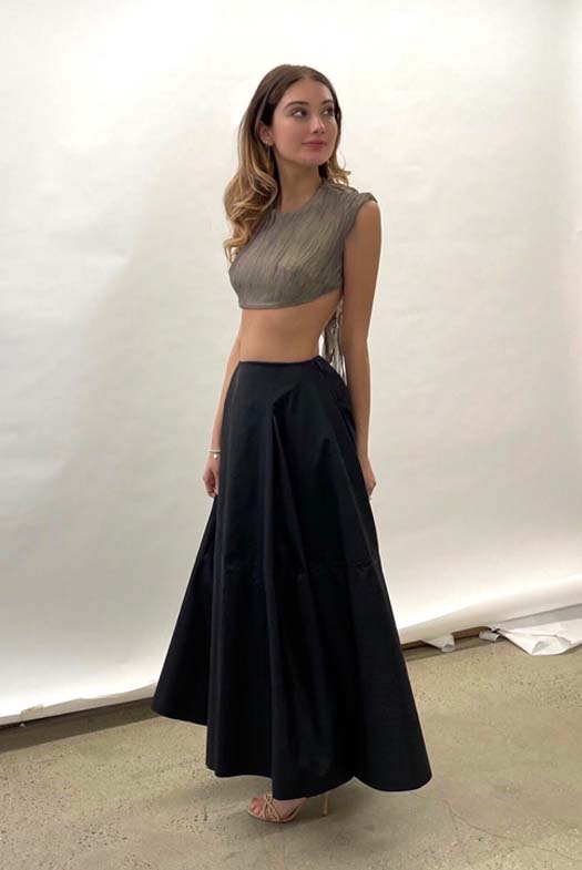 Girl leaning forward with her arms behind her back in a green silk-blend crop top that is textured. She is also wearing a floor-length pleated faux leather skirt.