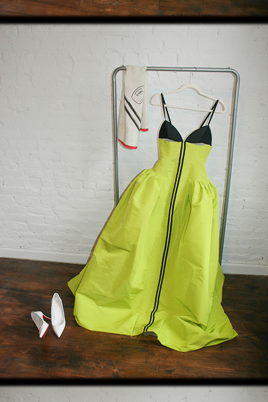 The Surfboard Dress hanging on a clothing rack against a white brick wall, with a silk headscarf hanging to the right and white patent leather pumps on the floor to the left.