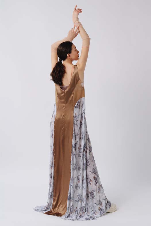 Back view of the dress showing the pleating from both sides and a draped scooped back. The model has her hands up in the air playing with the scrunched-up mesh arm warmers.