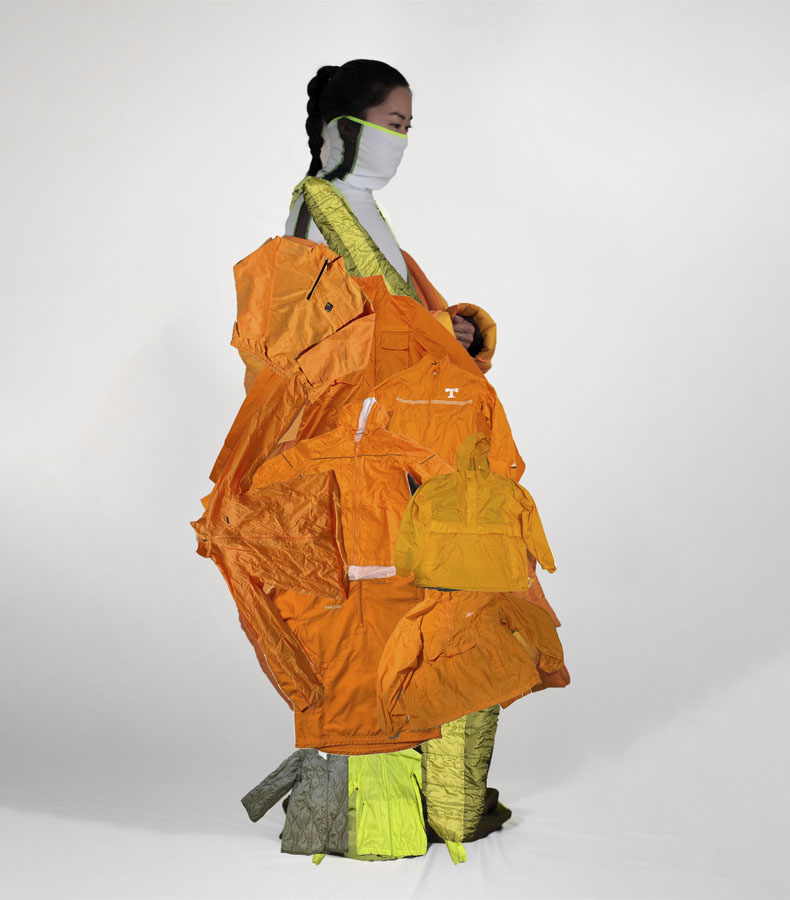This image shows a side view of a girl with black hair in the middle of a white background. She is wearing a white and green bodysuit that covers half of her face. She also wears a large orange jacket that is comprised of many smaller orange jackets. Below that, her pant legs are made from olive and neon green jackets.