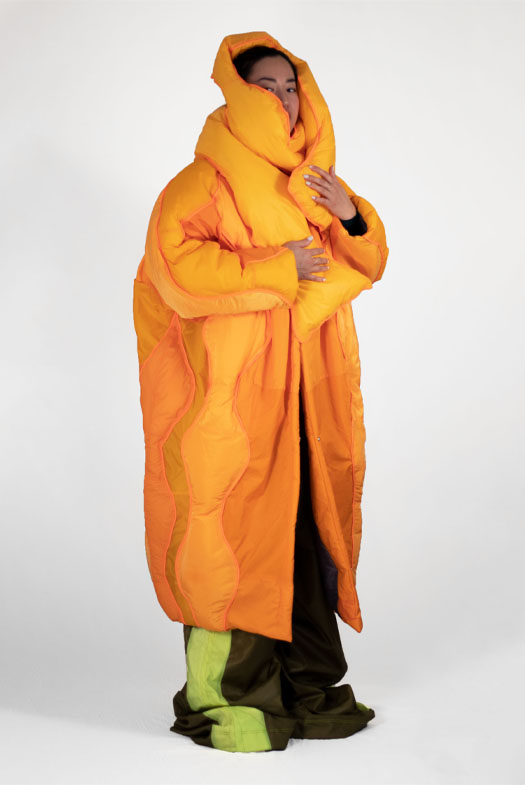 This image shows a 3/4 view of a girl with black hair in the middle of a white background. Most of her face is covered by a bright orange insulated scarf. She wears a large calf-length orange coat decorate with round, long pieces. Her pants show from the bottom of the coat and are a dark green with neon stripe.