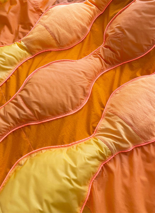 This image shows a detail shot of the orange jacket. There are different shades of orange shown in the round long pieces that are stuffed with insulation. Each piece is outlined by neon orange piping.