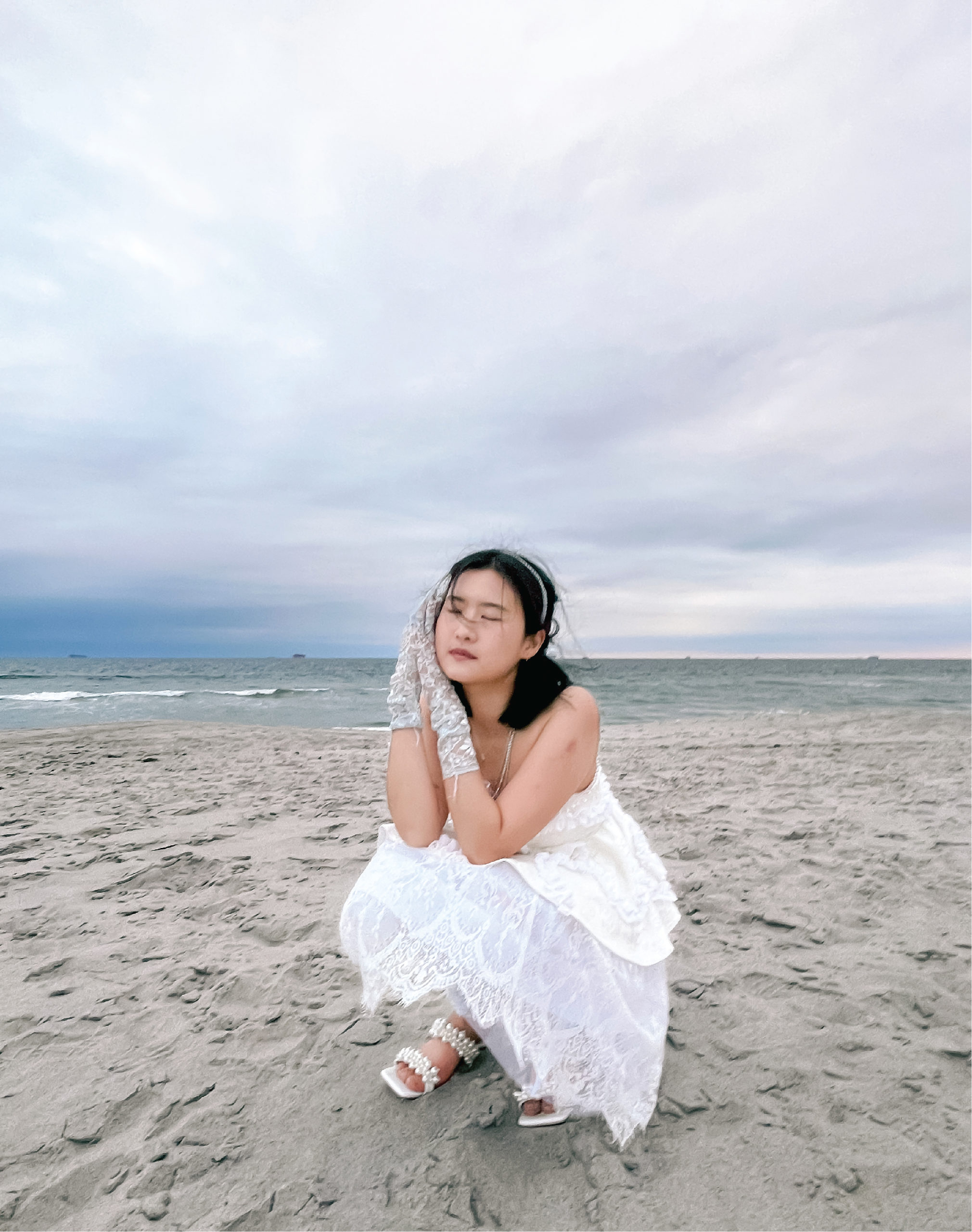 Model sits and places her hand against her head as if she is sleeping. She is wearing the strapless dress, pearl heels, and lace gloves. She is on the sand, and the water is in the background.
