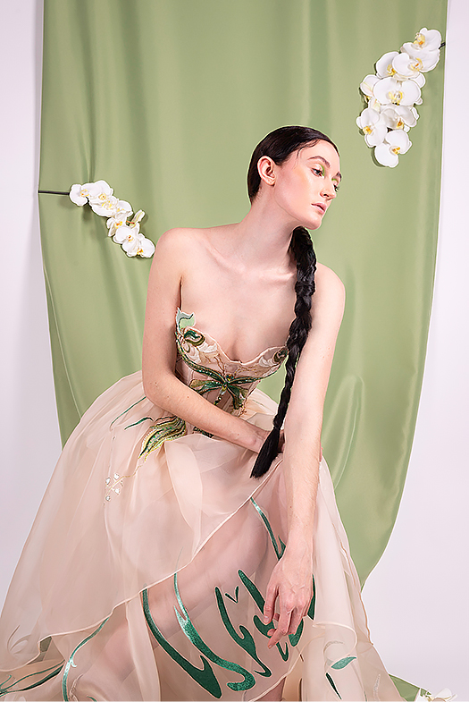 Half body shoot. Kiwi color fabric background with a little bit gathered, with three white orchids on top. The girl sits toward right in front of the kiwi fabric, with right hand on her knee and left hand toward front. Her long, dark brown braid falls over her left shoulder to her knee.