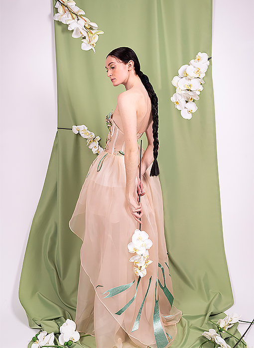 Full body shoot. Kiwi color fabric background with a little bit gathered, with three white orchids on top, gathered on the ground and covered with white orchids. The girl stands in the middle and toward the left with both hands placed on the back and hanging naturally, a long white orchid. Her long, dark brown braided hair hangs down to her waist. There are green thread embellishments like leaves around the hem.