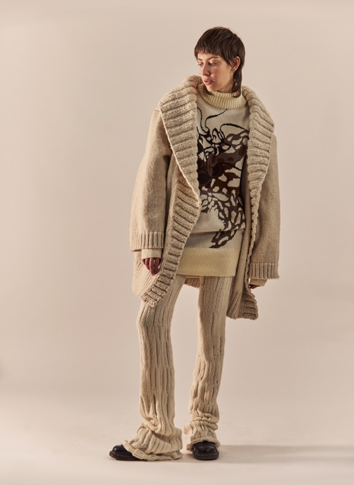 Front view of the whole body. The model has her body almost straight, showing the three layers of the look (pants, sweater, and cardigan). It has an oversized, relaxed look. The cardigan is open, showing the print of the sweater. The pants are scrunched at the bottom, on top of black loafers.