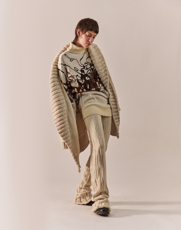 The model is leaning backwards with one leg in front of the other. The cardigan is falling off one of her shoulders, and her face is looking down. The light reflects on the pants, showing the pattern created by the travelling drop stitches.