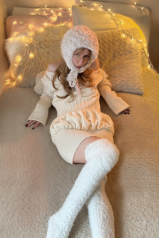 Photo of a girl sitting on a bed wearing ecru multi-stitch dress with white knit thigh-high socks and crocheted fur bonnet