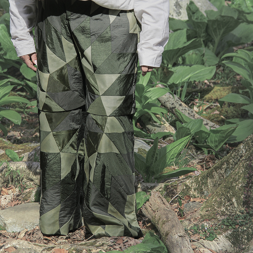 Person standing in the woods wearing a white short-sleeve shirt and green pants with a quilting pattern on them. The person is tying closed the strings that open and close the pockets on the top.
