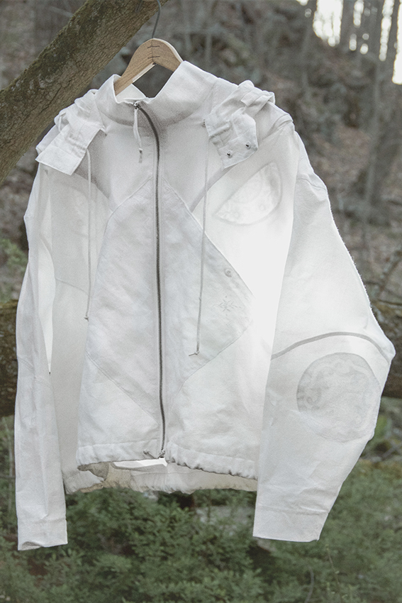 Person walking through a field of bright green skunk cabbage in the woods wearing the white cotton jacket with the white cotton tie pants. The tie pants look like large bandannas or squares tied together on the sides.