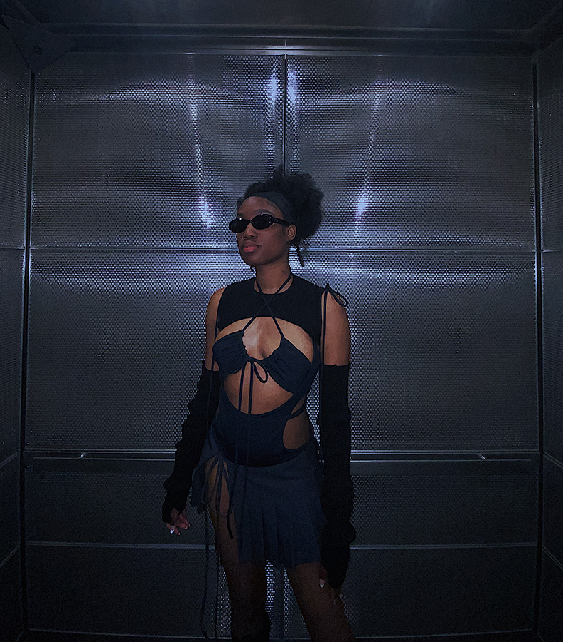A 3/4 view of the model. She is wearing black sunglasses and is standing in an elevator with silver metallic walls. She is wearing a super-cropped black knit top that sits above the bust and knit arm warmers. She is wearing a black bodysuit with cutouts on the side with a gathered halter bra underneath. She is wearing a low-rise dark gray pleated skirt with a slit on the right leg.