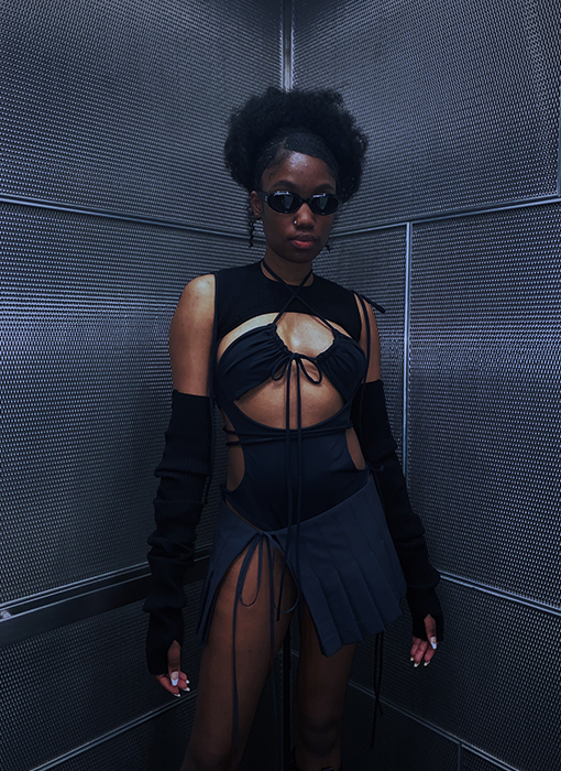 A close-up of the model. She is facing forward, her hip is swayed to the side, and her arms are flared to the side. She is wearing the same look. There is a clearer view of the straps tied around the waist of the bodysuit and the crossover strap detail of the halter bra.