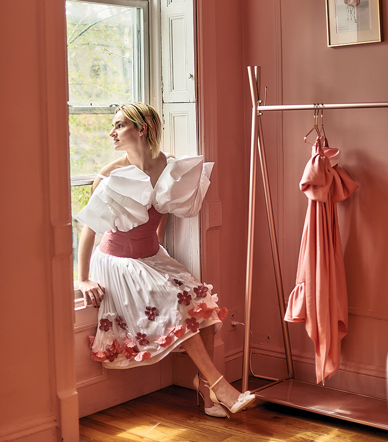 This image is of a model with blonde hair sitting in a windowsill looking out the window. The walls of the room are pink, and to the right of the model is a pink garment rack with a pink dress hanging on it. The model is wearing a garment that has a white 3D pleated satin structured puff-like bodice section that goes into a structured rose pink leather bodice with top stitching. The dress has a dropped waist, and attached to the leather bodice is a pleated white satin skirt with two layers. It is draped over the model's legs below her knee. The bottom of the skirt has flowers that are white, bright pink, and rose pink appliquéd on the hem of the skirt and flow upward to the waist. The model wears matching white satin shoes with a white flower on them. It is made of all sustainable fabrications: rose pink Piñatex leather made from pineapple leaves, white recycled polyester satin made from 100% post-consumer rPET recycled plastic water bottles, and white Tencel lining made from wood pulp. 