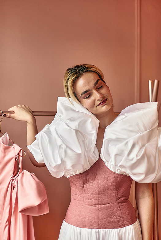 This image is of a model with her eyes closed. She has pink eye shadow and sparkly lipstick.  She is in front of a pink wall, and her left hand is on a pink garment rack with a pink dress hanging on it. She has blonde hair. She appears to be smiling, but her mouth is closed. The model is wearing a garment that has a white 3D pleated satin structured puff-like bodice section that goes into a structured rose pink leather bodice with top stitching. The dress has a dropped waist, and attached to the leather bodice is a pleated white satin skirt. The bodice white satin fluffy pleated sections cover her shoulders and the top part of her arms. It is made of all sustainable fabrications:  rose pink Piñatex leather made from pineapple leaves, white recycled polyester satin made from 100% post-consumer rPET recycled plastic water bottles, and white Tencel lining made from wood pulp. 