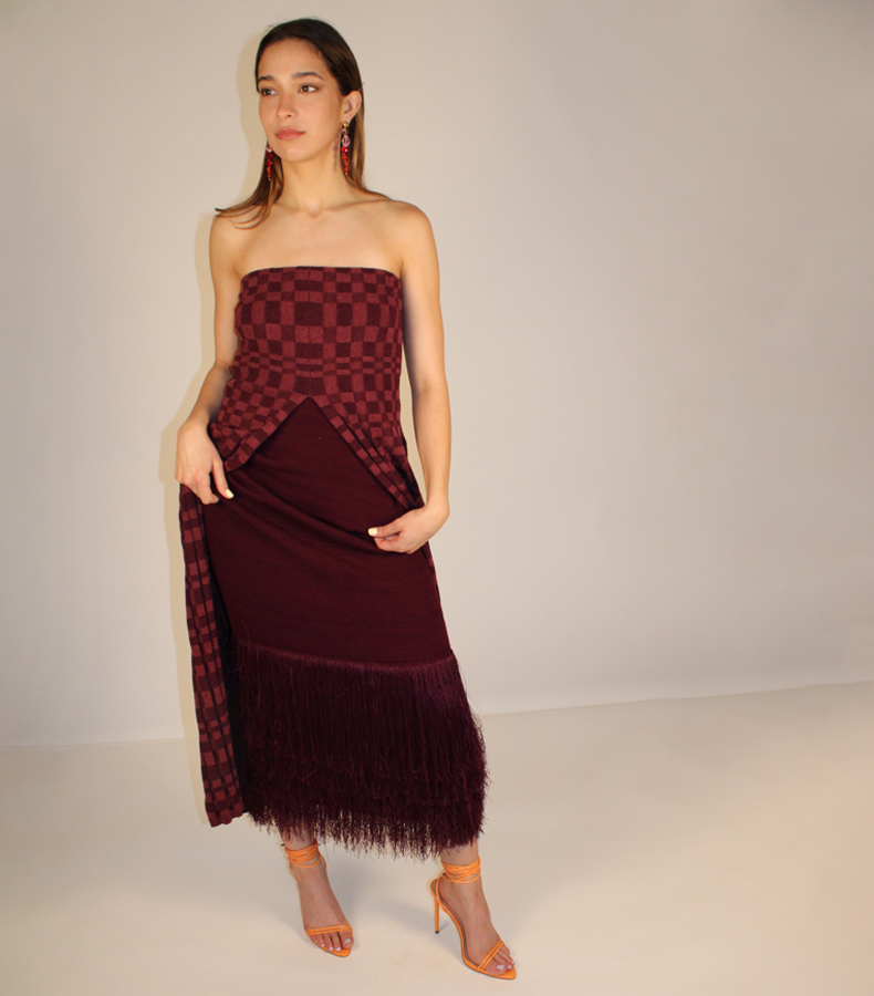 Model stands holding her dress up with two hands and looking off to one side. She wears a two-tone (burgundy and rose) plated rib strapless dress with a slit at center front and a burgundy dress with fringe underneath. She wears drop crystal earrings in a gradient from orange to pink to red. Her makeup is minimal and her nails are done. Her heels are orange with thin straps that wrap around the heel. She stands against a white background.