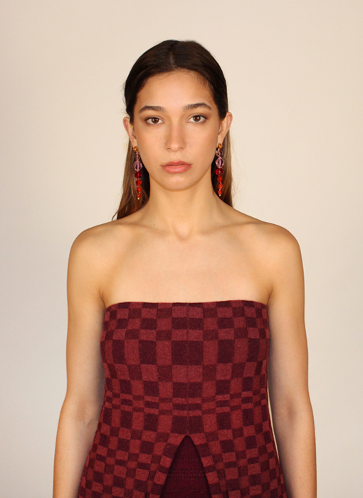 Portrait photo of model looking straightforward at camera against white background. Only waist up is visible. She has a straight face. She wears a two-tone (burgundy and rose) plated rib strapless dress with a checkered pattern and a slit at center front. She wears a burgundy garment underneath. Her hair has a center part and falls straight back. Her makeup is minimal and she wears drop crystal earrings in a gradient from orange to pink to red.