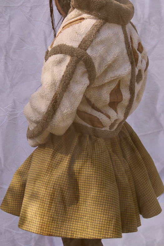 Image 3 is collaged showing a close-up photo of the back of the shearling coat. We see more of the full skirt and the brown trimmings on the cream shearling coat. Laser-cut details on the back in the shape of flowers.