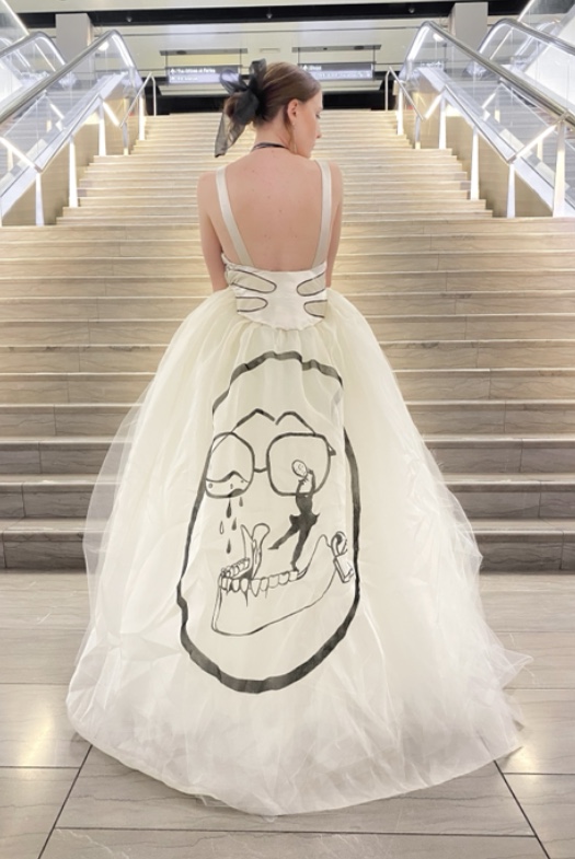 Model standing, view from the back. Hair tied in bun with a sheer black bow. Low, open back ivory dress. Beige leather patchwork edged in black. Circular black print on back of the dress.