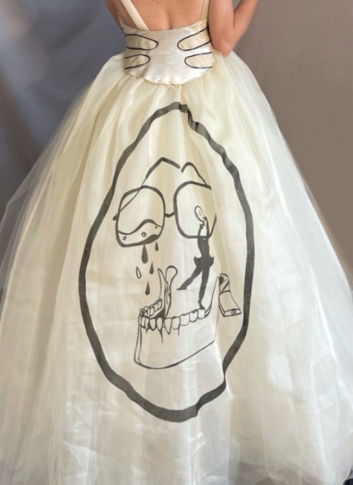 Model standing, view from the back. Hair tied in the bun with a sheer black bow. Low open back ivory dress. Beige leather patchwork edged in black. Circular black print on back of the dress.